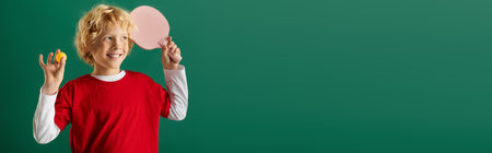 A cheerful boy with wavy blonde hair holds a ping pong set on a green backdrop, bannerの写真素材