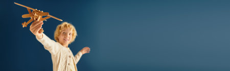 A happy blonde boy holds a wooden airplane while joyfully playing in a bright studio, bannerの写真素材