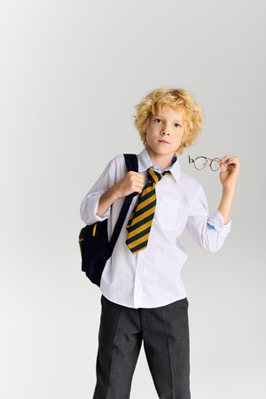 A young boy with wavy blonde hair stands confidently, holding his glasses and backpack.の写真素材