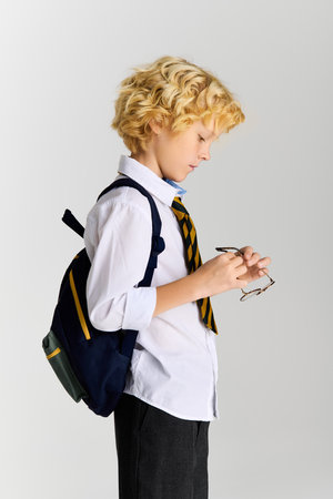 A young blonde boy with wavy hair is adjusting his glasses while wearing a school uniform.