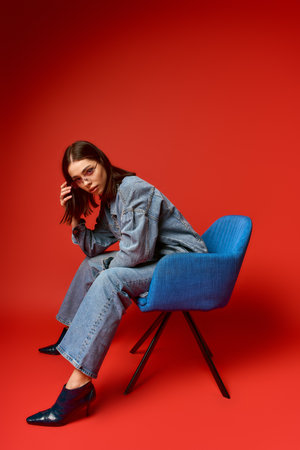 Young woman in a chic denim outfit poses confidently against a bold red backdrop.の写真素材