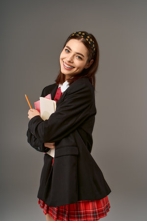 A cheerful young woman in a college uniform smiles brightly while holding her books in a studio.の写真素材