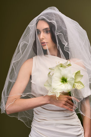 A young bride stands gracefully in a soft gown, her veil framing her face and flowers in hand.の写真素材