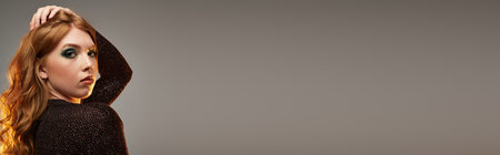 A stylish young woman with vibrant red hair showcases bold eye makeup in a chic grey studio, bannerの写真素材