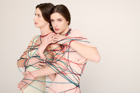Stylish twin sisters pose gracefully, intertwined with colorful strings in a bright studio space.の写真素材
