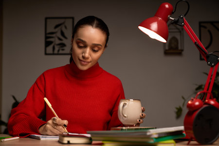 In her modern apartment, a young African American woman in a red turtleneck enjoys writing with tea.の写真素材