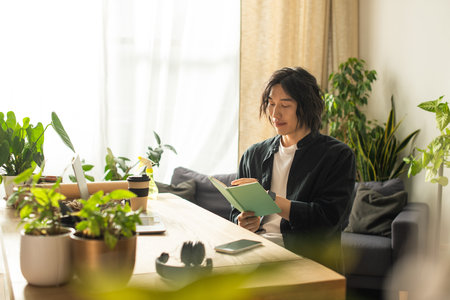 A young Asian florist writes in a notebook at a wooden table surrounded by vibrant plants.