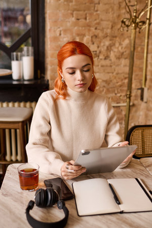 A young woman with red hair uses her tablet while sipping a warm drink in a cafe.