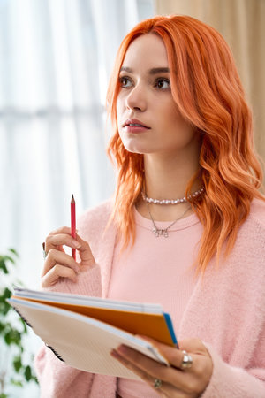 A young woman with red hair ponders creatively while holding colorful notebooks at home.