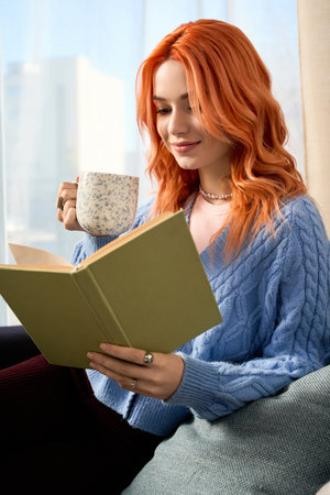 Young woman with red hair enjoys a warm drink while reading at home, surrounded by comfort.の写真素材