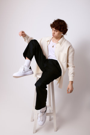 Young man in trendy outfit strikes a stylish pose on a stool, embodying modern fashion.の写真素材