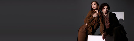 Young couple poses together in trendy outfits against a dark grey backdrop, banner