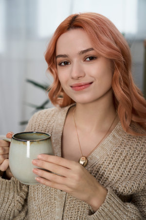 A young woman with soft pink hair smiles, holding a warm drink in a serene setting.の写真素材