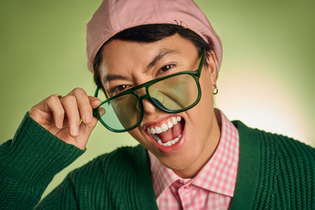 A fashionable asian man in a pink checkered shirt and cardigan strikes a fun pose in a green studio.の写真素材