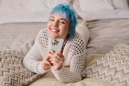 A vibrant young woman with blue hair relaxes on a bed, smiling while holding her phone.