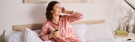 A lovely woman in pink pajamas relaxes on her bed, sipping coffee in her cozy home.