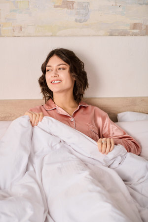 Sunlight fills the room as a lovely woman relaxes in bed, smiling under soft blankets.