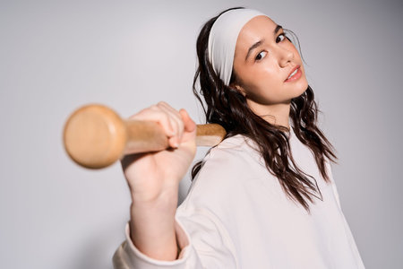 Young woman in sweatshirt exudes confidence while exercising with a wooden bat in a grey studio.
