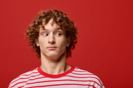 Stylish young man with curly hair wearing a striped t-shirt, posing thoughtfully in a red studio.