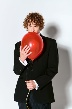 A joyful young man in a tuxedo holds a vibrant red balloon, celebrating in a bright studio.