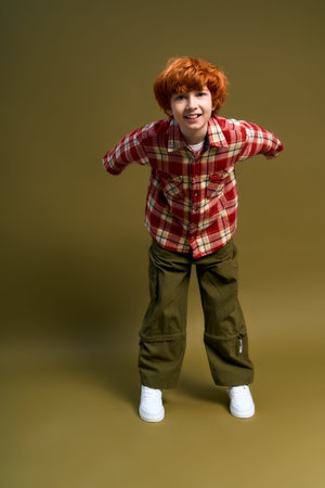 A little boy with vibrant ginger hair poses joyfully in a fashionable outfit, radiating style.