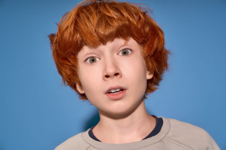 A stylish little boy with bright red hair expresses surprise, framed by a vivid blue backdrop.