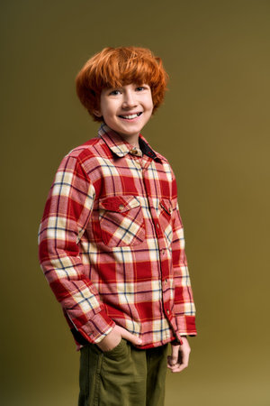 Charming boy with vibrant red hair smiles joyfully while displaying a stylish plaid outfit
