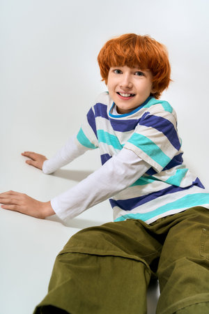 A fashionable young boy with bright orange hair smiles playfully while sitting on a white surface.