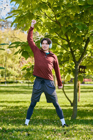 Curly-haired young man in a sport jacket strikes a happy pose under a leafy tree in spring.