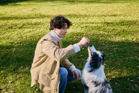 A young man with curly hair is joyfully training his Australian Shepherd in a lush park setting.