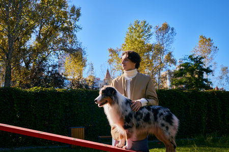 A young man in a spring jacket proudly training his Australian Shepherd under a clear blue sky.