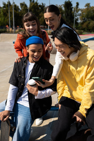 Four friends gather outdoors, sharing laughter while looking at a phone in a sunny skatepark.