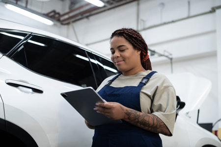 Young African American woman in overalls works on car repairs while using a tablet.の写真素材