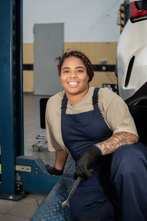Young woman in overalls joyfully repairs a car inside a bustling auto repair shop.