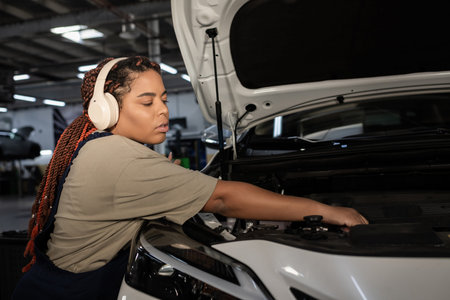Skilled Black woman in overalls and headphones works on car maintenance in a lively garage.
