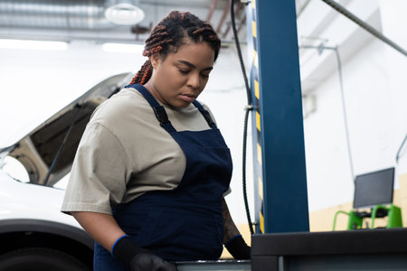 A young Black woman in overalls works on car maintenance in a sunny garage.