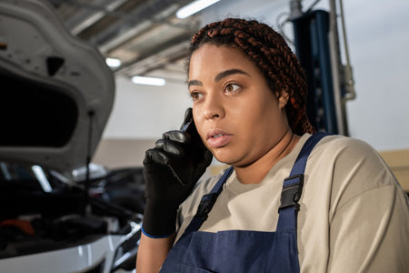 Dedicated African American woman in overalls talks on the phone while fixing a car engine