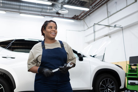 Young woman in overalls smiles while fixing a car in a busy repair garage.の写真素材