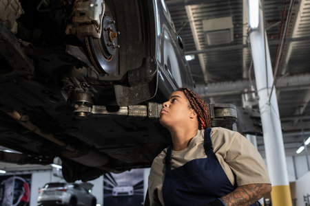 Young African American woman in overalls focuses on fixing a car in the garage environment.