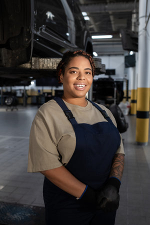 Joyful young woman in overalls proudly working on a car while smiling in a busy garageの写真素材