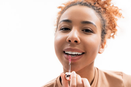 A young woman with curly hair smiles at the camera while applying lip gloss in a cozy studio.の写真素材