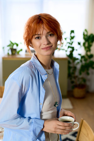 Smiling young woman with red hair holds a cup of coffee in a bright, leafy home setting.
