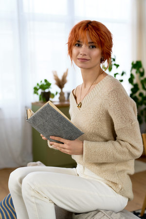 A young woman with bright red hair sits at home, cozy and immersed in her book.