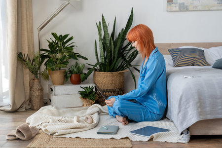 A young woman with vibrant red hair sits comfortably in her cozy bedroom, enjoying quiet time.の写真素材