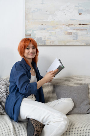 A cheerful young woman with red hair relaxes while reading a book in her modern living room.