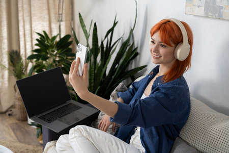 A young woman with striking red hair smiles while using her smartphone in a cozy living space.