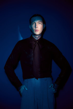 A young man stands confidently in stylish attire against a deep blue background.