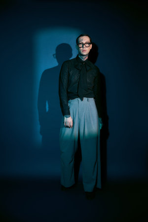 A young man stands confidently in a striking outfit, surrounded by deep blue shadows.
