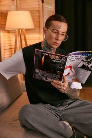 A young, fashionable man reads a magazine while relaxing on a stylish sofa in a warm setting.
