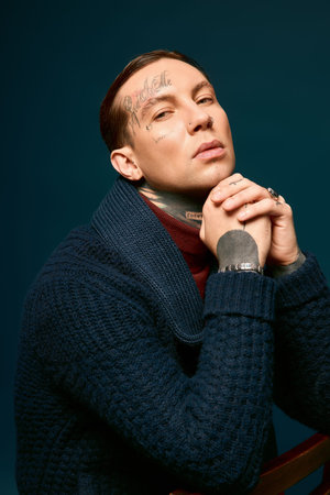 A stylish man rests his chin on folded hands, radiating charm and confidence.
